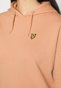 Sudadera con capucha color durazno de textura suave y un pequeño logo bordado en negro y amarillo de un pájaro en el pecho.
