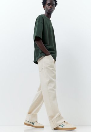 Man walking with hands in pockets wearing dark green oversized t-shirt, off-white wide pants, and white sneakers against plain white background.