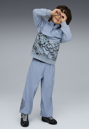 Niño vistiendo un sudadera gráfica azul claro y pantalones, posando con las manos enmarcando los ojos, de pie contra un fondo gradiente gris.