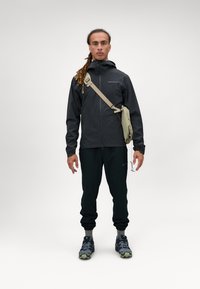 Sort vandtæt jakke med hætte, letvægtsstof, parret med sorte bukser og grå-grønne sko. Inkluderer en lille taske, der hænger over skulderen.