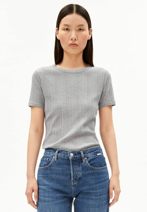 Femme aux cheveux raides et foncés portant un haut gris côtelé à manches courtes et un jean bleu taille haute sur un fond blanc uni.