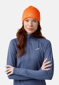 Beanie arancione con un design ricamato, indossato con una maglietta a maniche lunghe azzurro chiaro dotata di una zip a quarto e un piccolo logo sul petto.