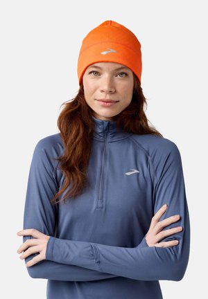 Oranje muts met een gestikt ontwerp, gedragen met een lichtblauw langmouwtop met een kwart rits en een klein logo op de borst.
