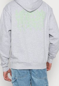 Felpa grigia con cappuccio regolabile. Presenta una grafica testuale in verde neon sul retro. Realizzata in un materiale morbido e di trama.