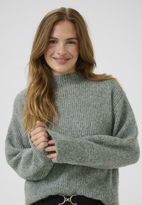 Jonge vrouw met lang golvend haar draagt een gezellige groene geribbelde trui, glimlachend terwijl ze met beide handen één mouw vasthoudt tegen een effen achtergrond.