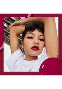 Modell med kort, mörkt lockigt hår som bär en vit t-shirt, med djärv burgunderläppstift och skarpa eyeliner mot en vinröd bakgrund.