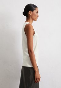 Helle beige ärmellose Bluse mit rundem Ausschnitt und glatter Textur, kombiniert mit einem schwarzen Lederrock, der ein Seitenprofil zeigt.