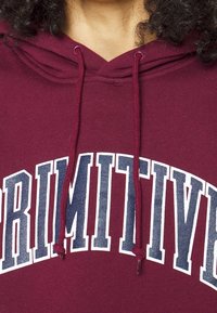 Gros plan sur un sweat à capuche bordeaux avec le texte "PRIMITIVE" en blanc et bleu marine sur la poitrine et des cordons pendants à l'avant.