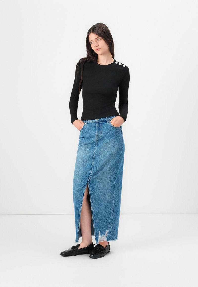 Kobieta w czarnej bluzce z długim rękawem i guzikami na ramionach, granatowej spódnicy jeansowej o wysokim stanie z przednim rozcięciem oraz czarnych loafersach, stojąca z rękami w kieszeniach.