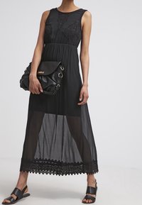 Svart maxiklänning med en figursydd bodice och spetsdetaljer, som har en genomskinlig kjol och scallopad spetskant. Matchad med en svart axelväska.