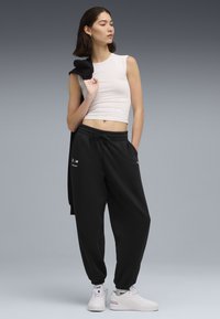 Sorte joggingbukser med elastiske manchetter og træksnor i taljen, parret med en tætsiddende hvid tanktop. Modellen holder en sort jakke over den ene skulder.