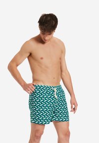 Costumi da bagno da uomo in tessuto verde teal con un motivo geometrico bianco, dotati di una vita elastica e di un cordoncino per la regolazione.