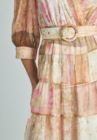 Gestufte Kleid mit durchscheinendem, leichtem Stoff in Pink- und Beigetönen. Details umfassen einen breiten Gürtel mit einer runden Schnalle und floralen Mustern.