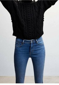 Suéter negro de punto trenzado con cuello redondeado, combinado con jeans ajustados azul de tiro medio que presentan un diseño de cinco bolsillos y un ligero desgaste.
