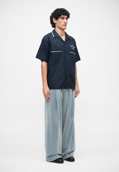 Chemise marine à manches courtes avec des accents blancs et une broderie sur la poitrine, associée à un pantalon large gris clair et des chaussures noires.