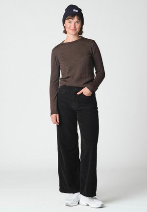 Jeune femme portant une chemise marron foncé à manches longues, un pantalon large noir, des baskets blanches et un bonnet noir, debout avec une main dans la poche.