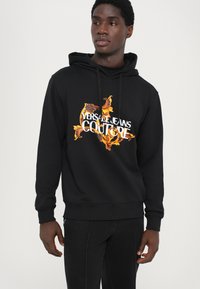 Črna kapuca s puloverjem, ki ima v središču pisano logotipno zasnovo "Versace Jeans Couture" v oblikovanju plamena, z rebrastimi manšetami in robom.