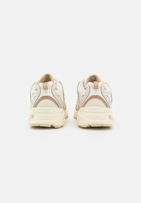 Sneakers bianchi con una tomaia in rete testurizzata, dettagli beige e una suola ammortizzata. Presentano un design curvo e un collare imbottito per il comfort.