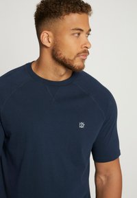 Camiseta de manga corta azul marino, hecha de material suave, con mangas raglán y un pequeño logo blanco en el pecho. Textura suave.
