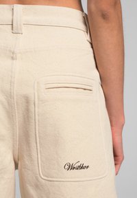 Pantaloni in cotone beige con una tasca posteriore e logo ricamato "Wrstbhvr" marrone. Tessuto textured con dettagli delle cuciture visibili.