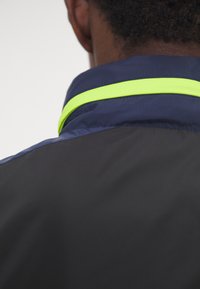 Navy windbreaker jacket with a contrasting reflective yellow collar. Features smooth texture and sleek design, emphasizing functionality.