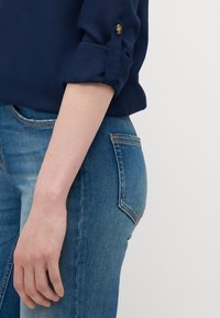 Blouse bleu marine avec manches retroussées et détail de boutons dorés, associée à un jean ajusté bleu clair avec une poche arrière et un léger dégradé.