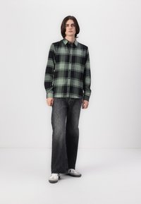 Camisa de cuadros verde y negra con botones combinada con jeans negros desgastados. El modelo está de pie con las manos en los bolsillos, vistiendo zapatillas blancas.