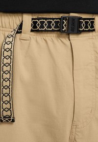 Beżowe szorty cargo z elastycznym paskiem w talii, wyposażone w teksturowany pas w czarno-beżowy wzór oraz czarną klamrę. Prosty przód i boczne kieszenie.