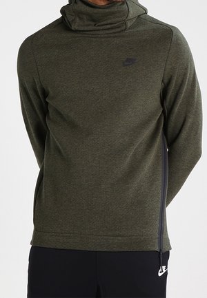 Mężczyzna noszący oliwkowo-zieloną bluzę Nike z kapturem i wysokim kołnierzem oraz czarne spodnie Nike, stoi z rękami za plecami.