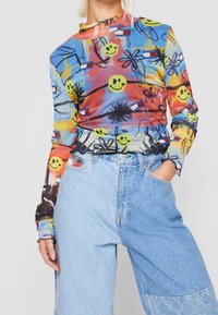 Long-sleeve shirt in a multicolored design featuring smiley faces and floral patterns, made of lightweight fabric, paired with light blue oversized jeans.