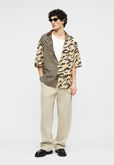 Άνδρας που φοράει μαύρα γυαλιά ηλίου, μπεζ παντελόνι, λευκό μπλουζάκι και κοντομάνικο πουκάμισο με καρό και animal print μοτίβα.