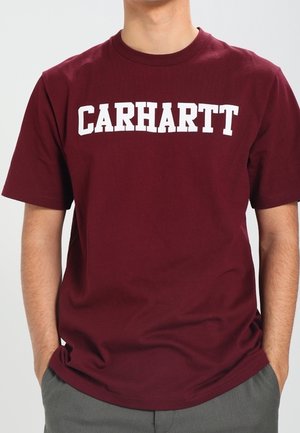 Camiseta de manga corta color granate con el texto blanco "CARHARTT" en el pecho, llevada por una persona con las manos en los bolsillos de unos pantalones grises.