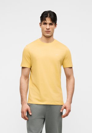 Νεαρός άνδρας στέκεται, φοράει κίτρινη κοντομάνικη μπλούζα και γκρι παντελόνι, κοιτάζοντας προς τα εμπρός σε απλό ανοιχτό φόντο.