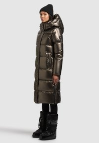 Fémes sötétzöld, kapucnis kabát steppelt mintával és oldalzsebekkel, fekete bélelt csizmával párosítva.