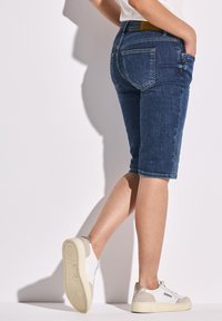 Dżinsowe szorty bermudy w ciemnoniebieskim kolorze, o średnim stanie z pięcioma kieszeniami. Połączone z białymi i beżowymi sneakersami. Gładka tekstura, swobodny fason.