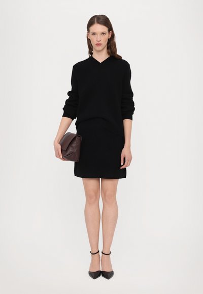 Schwarzer Pullover mit V-Ausschnitt, kombiniert mit einem schwarzen Rock. Eine dunkelbraune Lederhandtasche wird gehalten, die ein gefaltetes Klappen-Design aufweist. Schwarze spitze High Heels getragen.