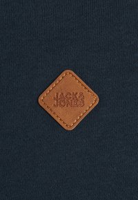 Feltro di pelle a forma di diamante con il testo in rilievo "JACK & JONES" cucito su un tessuto lavorato a maglia blu scuro.