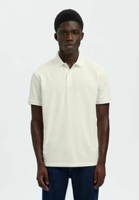 Jeune homme à la peau foncée portant un polo blanc uni à manches courtes et un jean bleu foncé, debout devant un fond clair.