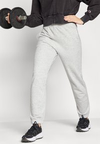 Grå bomullssweatpants med elastiske mansjetter, sammen med svarte treningssko. Modellen holder en svart manual i en sporty stilling.