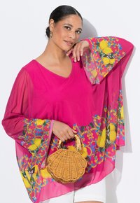 Blusa rosa semi-trasparente con stampa colorata di limoni sulle maniche, abbinata a una borsa rotonda in rattan intrecciato. La modella posa su uno sfondo bianco.