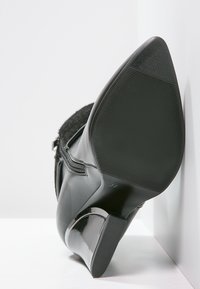 Bota negra de tacón alto con un acabado de charol brillante y parte superior texturizada. Cuenta con una suela de goma con un patrón acanalado para mayor agarre.