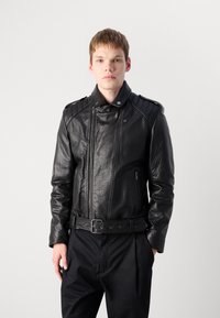 Young man with light skin and short brown hair wearing a zipped black leather jacket and black pants against a plain light background.