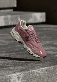 Ροζ και λευκό αθλητικό παπούτσι New Balance 1000 κρεμασμένο στον αέρα πάνω από σκάλες από σκυρόδεμα, με σκιά ορατή από κάτω.