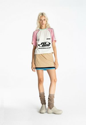 Joven mujer con cabello rubio que lleva una camiseta gráfica blanca y rosa, mini falda beige con ribete azul, calcetines beige caídos y zapatos tipo slip-on texturizados.