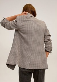 Personne aux cheveux longs portant un blazer beige et noir à carreaux et un jean noir, se grattant la nuque, dos tourné vers la caméra.