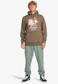 Brown cotton hoodie with logo and graphic print, paired with light green sweatpants. Model wears black high-top shoes.