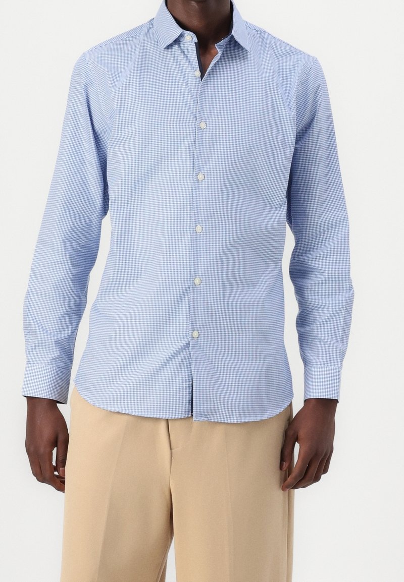 Homme portant une chemise à manches longues à petits carreaux bleu clair et blanc avec un pantalon beige, debout devant un fond uni.