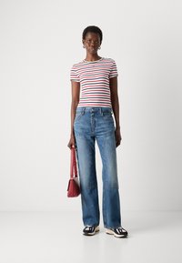 T-shirt rayé rouge et noir associé à un jean large délavé, baskets noire et blanche, tenant un sac à main rouge.