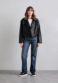 Schwarze Fake-Leder kurze Jacke mit breiten Revers, getragen über einem weißen Oberteil. Blaue gerade Jeans und schwarze Sneaker mit weißen Sohlen.