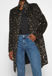 Djurprintad kappa i mörkbruna och svarta toner, med hög krage, texturerat tyg och svarta knappar, kombinerad med ljusblå jeans.